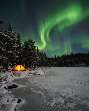 Green aurora over a frozen lake with snowy pines, a single tent glowing warm at the shore, no people. With the Chrome Film preset applied.