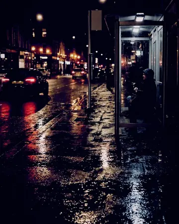 A bus stop on a wet pavement, reflections in the puddles, night, lit only by street and shop lights. With the Cinema Grade preset applied.