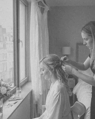 A fictional bride having her hair pinned by a window in a hotel room, white dress hanging behind, soft daylight. With the Mono Film preset applied.