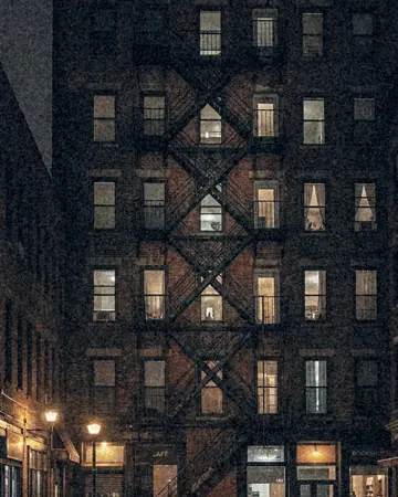 A fire escape zigzagging up a brick facade, night, lit only by street and shop lights. With the Night Halation preset applied.