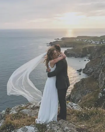 A fictional eloping couple embracing on a sea cliff at sunset, her veil blowing, warm backlight. With the Pastel Film preset applied.