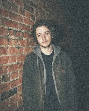 A fictional young man against a brick wall at night, direct camera flash, everything behind him falling to black. With the Pastel Film preset applied.