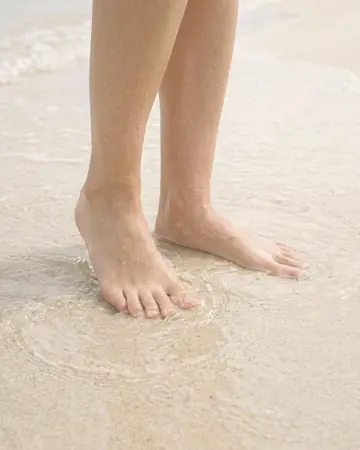 Bare feet in shallow clear water on pale sand, daylight. With the Warm Negative preset applied.