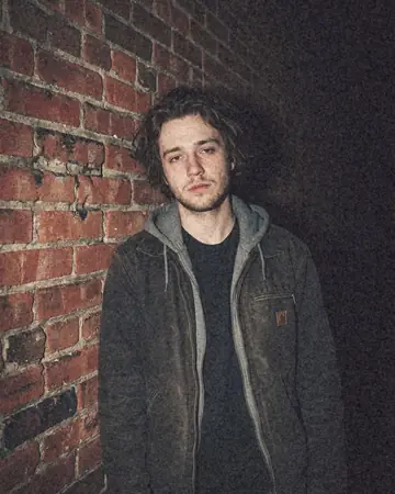 A fictional young man against a brick wall at night, direct camera flash, everything behind him falling to black. With the Warm Negative preset applied.