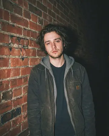 A fictional young man against a brick wall at night, direct camera flash, everything behind him falling to black. With the Warm Negative preset applied.
