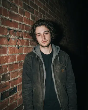 A fictional young man against a brick wall at night, direct camera flash, everything behind him falling to black. Photographed without a preset.