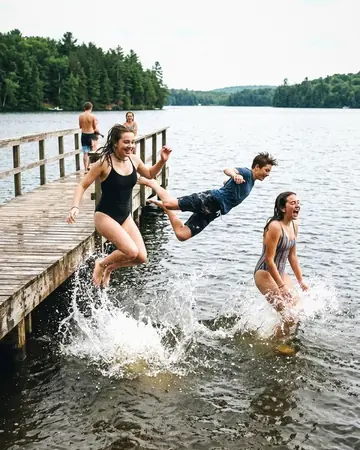 Fictional teenagers mid-jump off a wooden lake pier into the water, summer afternoon, splashes. With the Wedding Film preset applied.