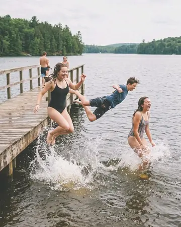 Fictional teenagers mid-jump off a wooden lake pier into the water, summer afternoon, splashes. With the Wedding Film preset applied.